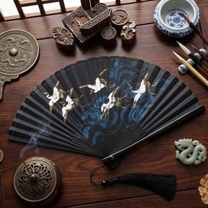 May include: A black folding fan with a crane design, featuring white birds and blue wave patterns. The fan is open, displaying its full design. A black tassel hangs from the base. The image also includes other Asian-inspired decor.