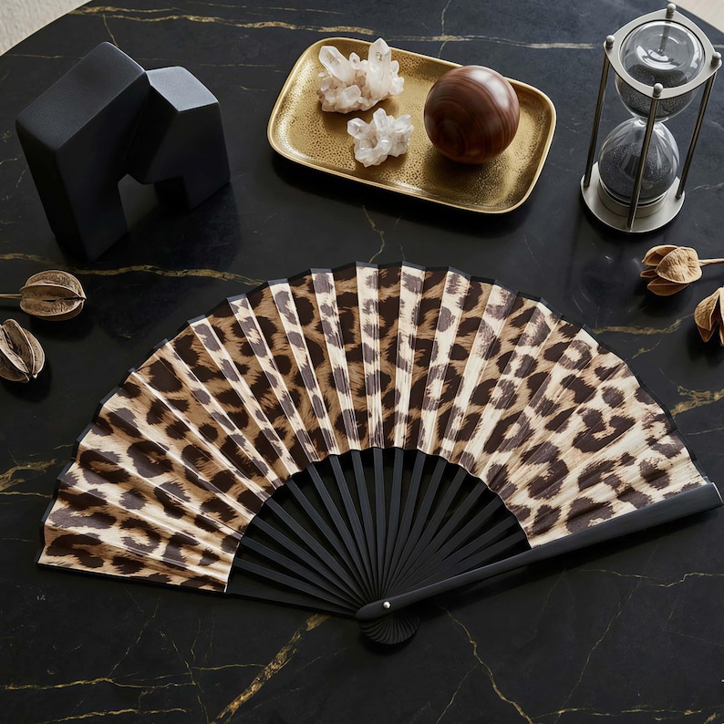 May include: An open hand fan with a black frame and a leopard print pattern. The fan is displayed on a black marble surface, alongside decorative items. The fan's design features brown and beige animal print.