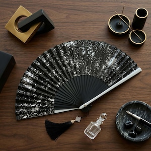 May include: A black hand fan with silver accents, a black tassel, and a small glass perfume bottle are displayed on a wooden surface. Other items include a gold and black sculpture, black and gold candle holders, and a marble dish with a pen.