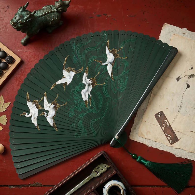 May include: A dark green folding fan with a crane design. The fan features white and brown cranes in flight, with green wave-like patterns. A green tassel hangs from the base. The fan is displayed on a red surface with other decorative items.