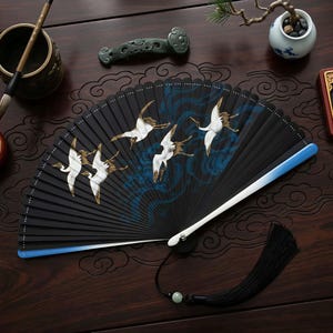 May include: A black folding fan with a crane design. The fan features white cranes with gold accents against a backdrop of blue waves. The handle is white and blue, with a black tassel and a jade bead.