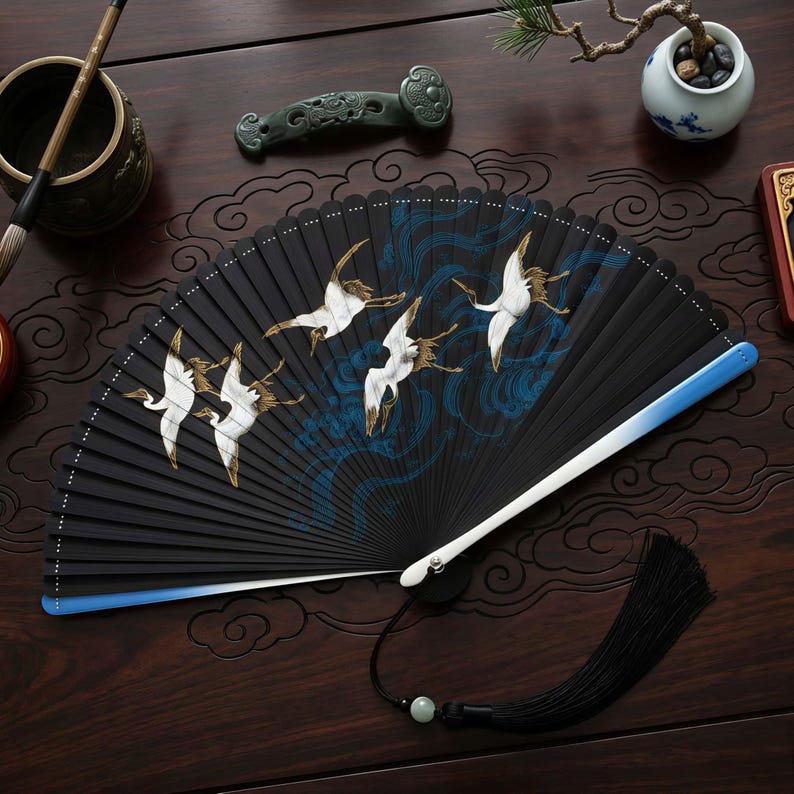 May include: A black folding fan with a crane design. The fan features white cranes with gold accents against a backdrop of blue waves. The handle is white and blue, with a black tassel and a jade bead.