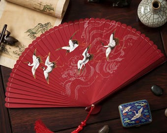 May include: A red hand fan with a crane design. The fan is open, displaying the detailed artwork of white cranes in flight against a backdrop of stylized clouds. A red tassel hangs from the base. The fan is on a dark wooden surface.