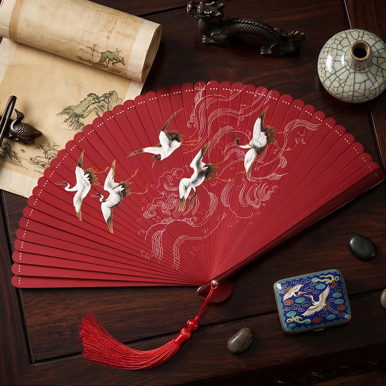 May include: A red hand fan featuring a crane design. The fan is open, revealing detailed artwork of white cranes in flight against a backdrop of stylized clouds. A red tassel hangs from the base. The fan is on a dark wooden surface.
