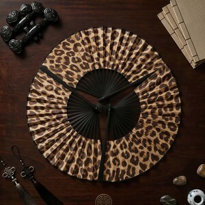 May include: Three black-handled hand fans with a leopard print design are arranged on a dark wooden surface. Decorative black objects and small stones are placed around the fans. The fans are open, displaying the animal print.