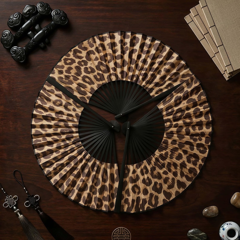 May include: Three black-handled hand fans with a leopard print design are arranged on a dark wooden surface. Decorative black objects and small stones are placed around the fans. The fans are open, displaying the animal print.