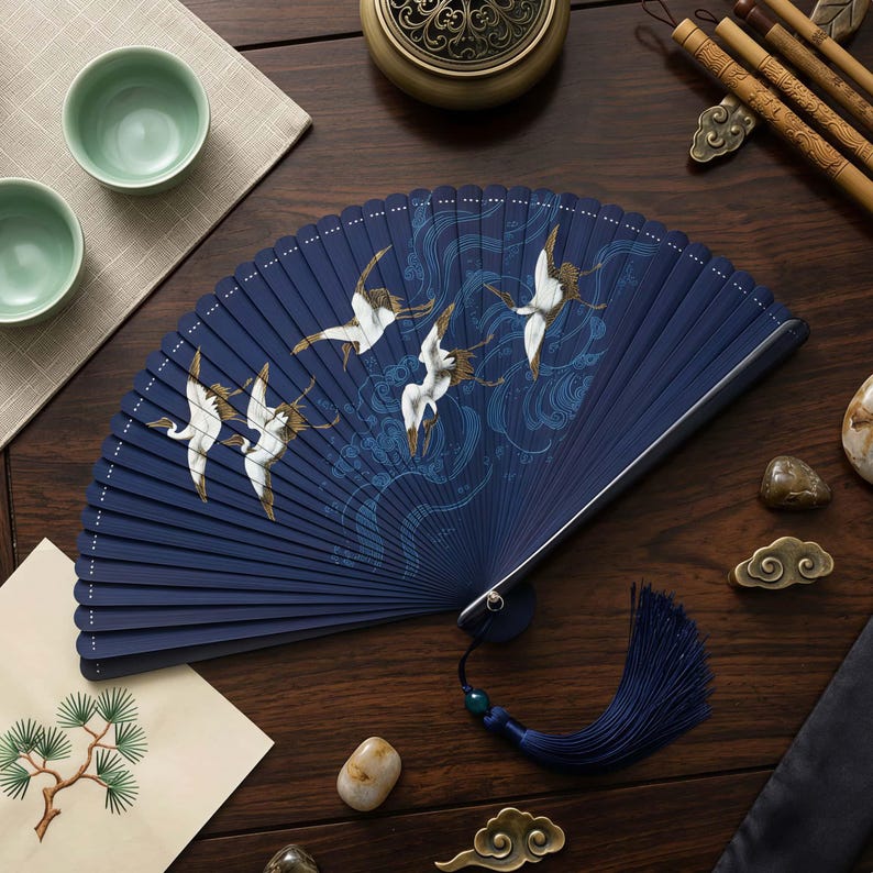 May include: A navy blue folding fan with a crane design. The fan features white cranes with gold accents against a background of stylized waves. A navy tassel hangs from the base. The fan is displayed on a dark wooden surface.
