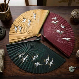 May include: Three hand fans in yellow, burgundy, and dark green, each featuring a painted design of flying birds. The fans are displayed on a dark wooden surface, highlighting their intricate details and colours.