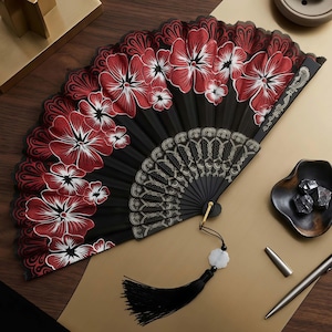 May include: A black hand fan with red and white floral designs. The fan has a black tassel with a white charm. The fan is open, revealing a detailed pattern on the lower portion. The fan is on a gold surface.