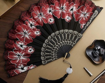 May include: A black hand fan with red and white floral designs. The fan has a black tassel with a white charm. The fan is open, revealing a detailed pattern on the lower portion. The fan is on a gold surface.