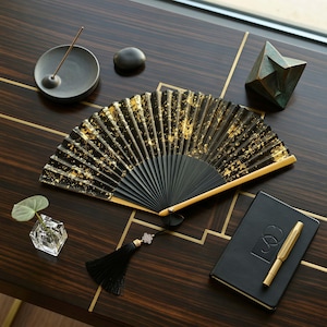 May include: A black hand fan with gold leaf accents is open on a dark wood surface. A black tassel with a decorative knot hangs from the fan. Other items on the surface include a notebook, pen, incense burner, and a small geometric sculpture.
