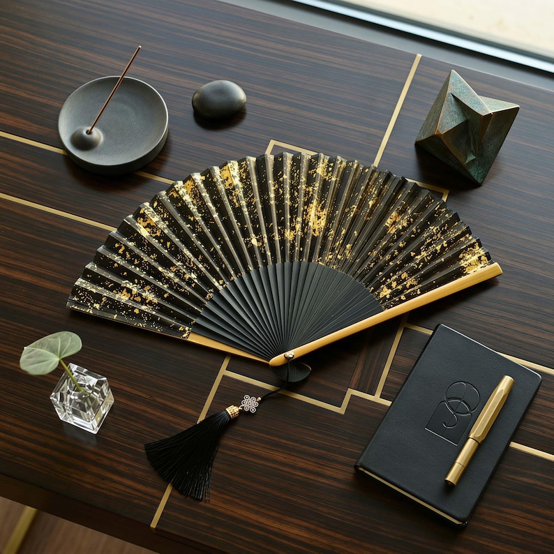 May include: A black hand fan with gold leaf accents is open on a dark wood surface. A black tassel with a decorative knot hangs from the fan. Other items on the surface include a notebook, pen, incense burner, and a small geometric sculpture.