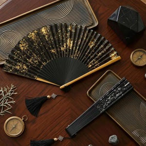 May include: A black hand fan with gold leaf accents, open and displayed on a wooden surface. A closed fan, trays, compasses, and a decorative black geometric object are also present. The fan has a black tassel.