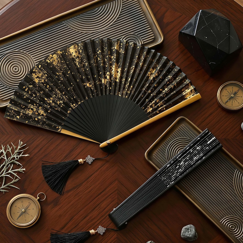 May include: A black hand fan with gold leaf accents, open and displayed on a wooden surface. A closed fan, trays, compasses, and a decorative black geometric object are also present. The fan has a black tassel.