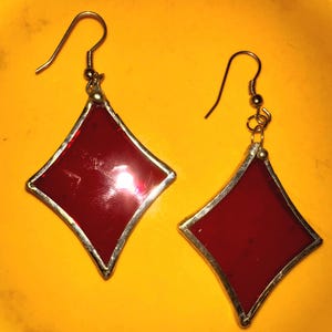 May include: A pair of red diamond-shaped earrings with a silver-colored border and gold-colored ear wires. The earrings have a shiny, reflective surface and are set against a yellow background.