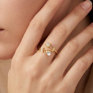 May include: A gold-colored ring featuring a floral design. The ring has a flower with a clear gemstone and a smaller flower with multiple small gemstones. The ring also has leaf accents.