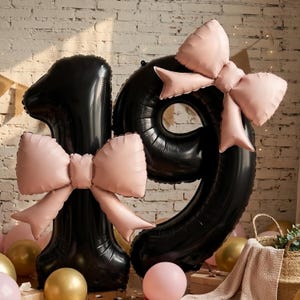 May include: Large black number balloons shaped as 1 and 9, each decorated with a soft pink foil bow. The number 1 features a bow at its center, while the number 9 has a bow at the top. These balloons are displayed alongside smaller round balloons.