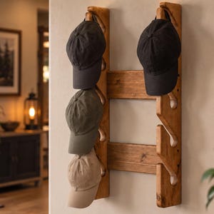 May include: A wall-mounted wooden hat rack featuring two vertical planks connected by two horizontal bars. The rack has four angled pegs on the left side and four on the right, currently holding three baseball caps in black, olive green, and tan.