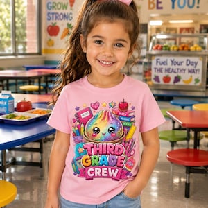 May include: A light pink t-shirt featuring a colorful graphic of a smiling, rainbow-colored dumpling surrounded by school supplies like books, pencils, and an apple, with the text "THIRD GRADE CREW" printed in bold, glittery pink letters.