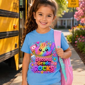 May include: A light blue t-shirt featuring a colorful graphic of a smiling, rainbow-colored character in a basket surrounded by school supplies like glue, scissors, and a notebook, with the text Second Grade ROCKS printed in bold, vibrant letters.