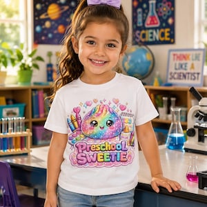 May include: A white short-sleeved t-shirt featuring a colorful graphic of a smiling, rainbow-colored dumpling surrounded by art supplies, including crayons, a paint palette, and glue, with the text "Preschool SWEETIE" printed in a playful font.