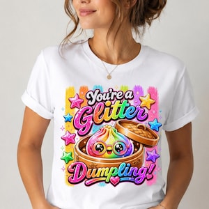 May include: A white t-shirt featuring a colorful graphic of a rainbow-colored dumpling with large eyes and glasses inside a bamboo steamer. The text reads "You're a Glitter Dumpling!" surrounded by vibrant, multi-colored stars and brush strokes.