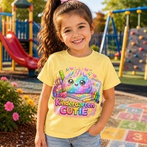 May include: A light yellow short-sleeve t-shirt featuring a colorful graphic of a smiling, rainbow-colored dumpling character surrounded by school supplies like crayons, a notebook, and an apple, with the text Kindergarten CUTIE.