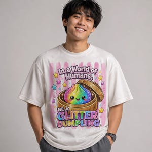 May include: A white short-sleeve t-shirt featuring a graphic of a rainbow-colored dumpling in a bamboo steamer. The text reads In a World of Humans, BE A GLITTER DUMPLING. The design includes pink vertical stripes and colorful stars.