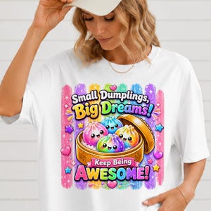 May include: A white t-shirt featuring a colorful graphic of three smiling, rainbow-colored dumplings inside a wooden steamer basket. The text reads "Small Dumplings, Big Dreams!" and "Keep Being AWESOME!" surrounded by stars, hearts, and vertical brush strokes.