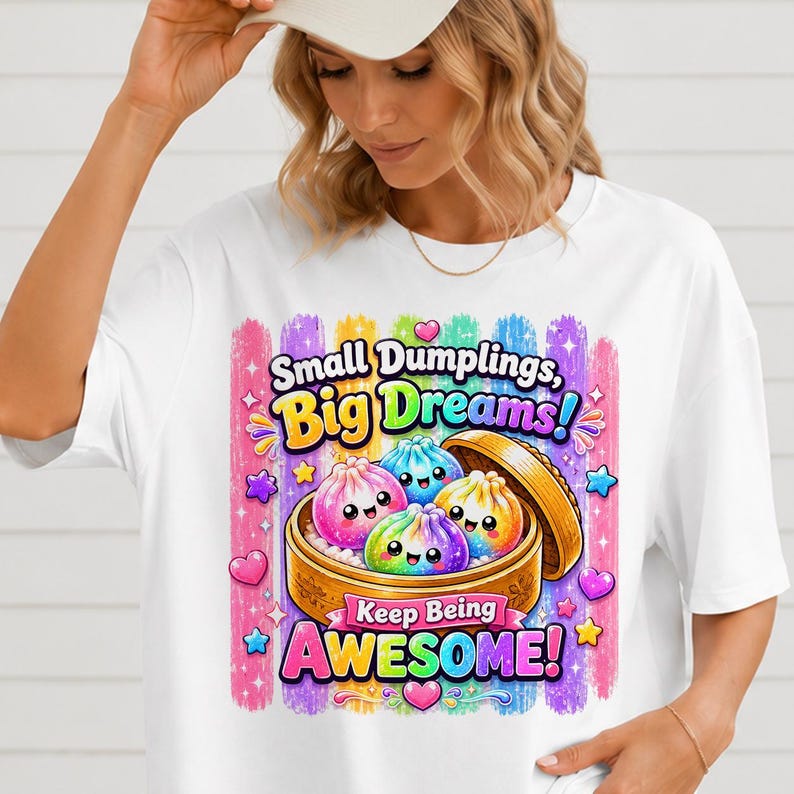 May include: A white t-shirt featuring a colorful graphic of three smiling, rainbow-colored dumplings inside a wooden steamer basket. The text reads "Small Dumplings, Big Dreams!" and "Keep Being AWESOME!" surrounded by stars, hearts, and vertical brush strokes.