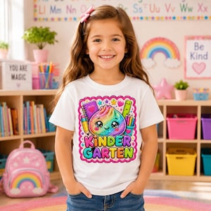 May include: A white t-shirt featuring a colorful graphic of a glittery, smiling dumpling character surrounded by school supplies like a notebook, pencil, and ruler, with the text "KINDER GARTEN" printed in bold, multi-colored bubble letters.
