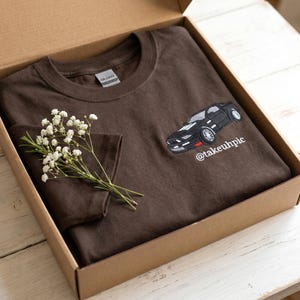 Może przedstawiać: Brązowa koszulka z czarnym haftem samochodu z przodu, prezentowana w kartonowym pudełku. Projekt samochodu zawiera białe i czerwone akcenty, a pod nim znajduje się napis "@takeuhpic". Gałązka białych kwiatów spoczywa na koszulce.