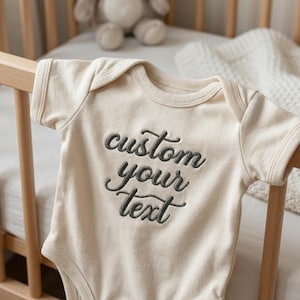 Puede incluir: Body de bebé color crema con el texto bordado "custom your text" en escritura gris oscuro. El body se muestra en una barandilla de cuna de madera, con un peluche y una manta en el fondo.