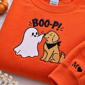 May include: An orange sweatshirt featuring an embroidered design of a white ghost touching the nose of a golden dog wearing a black bandana. Above the figures, the text reads BOO-P! in black. The sleeve cuff displays the letter M and a heart.