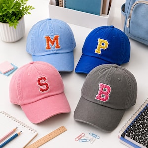 Puede incluir: Cuatro gorras de béisbol en azul claro, azul real, rosa y gris carbón. Cada gorra presenta en la parte frontal una letra grande de estilo universitario con purpurina: una M naranja, una P amarilla, una S roja y una B rosa, con borde blanco.