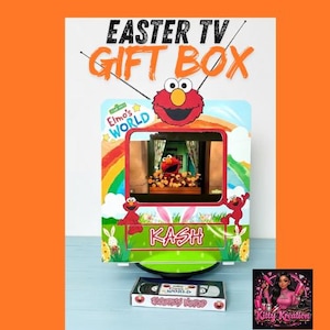 May include: A colorful Easter-themed gift box shaped like a television, featuring Elmo from Elmo's World. The box has a screen displaying Elmo, with the name "KASH" and a VHS tape. The words "EASTER TV GIFT BOX" are at the top.