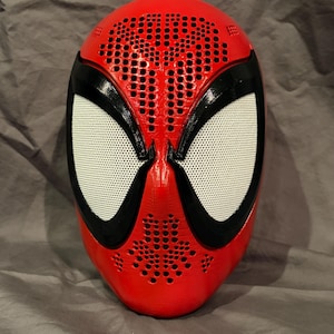 May include: A red full-face mask featuring a textured pattern of small circular indentations across the forehead and chin area. The mask includes two large, white, mesh-covered eye lenses framed by thick, glossy black borders that extend outward.