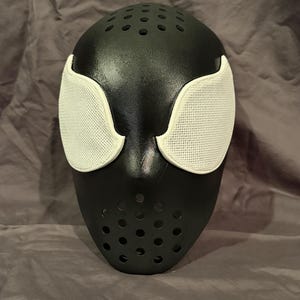 May include: A black full-face mask featuring a smooth, matte finish with a series of small circular ventilation holes located on the forehead and chin areas. The mask includes two large, almond-shaped white eye lenses with a fine mesh texture.