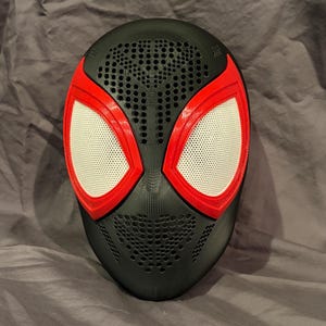 Può includere: Una maschera integrale nera caratterizzata da una superficie testurizzata e perforata sulla fronte e sul mento. La maschera presenta due grandi aperture oculari a mandorla bordate di rosso brillante con lenti a rete bianche.