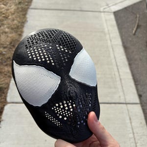 Könnte beinhalten: Eine schwarz-weiße Spider-Man-Maske mit strukturierter Oberfläche. Die Maske hat weiße Augenabdeckungen und ein perforiertes Design auf Stirn und Wangen. Die Maske wird vor einem grauen Bürgersteig gehalten.