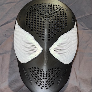 May include: A matte black full-face mask featuring a textured surface with numerous small circular ventilation holes across the forehead and mouth areas. The mask includes two large, almond-shaped white mesh eye lenses with a distinct border.