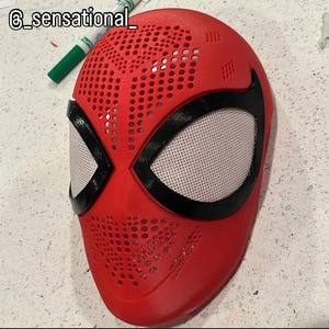 May include: A red mask featuring a textured surface with numerous small circular perforations across the forehead and chin areas. The mask includes two large, almond-shaped eye openings outlined in black with white mesh lenses. Text reads @_sensational_.
