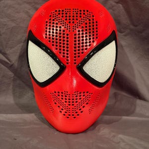 May include: A bright red full-face mask featuring a perforated texture across the forehead and mouth area. The mask includes two large, almond-shaped eye openings framed in black with white mesh lenses, creating a distinct superhero-inspired look.