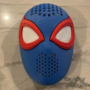 May include: A blue mask with red trim around the eyes, resembling a superhero mask. The eye openings are white mesh. The mask has multiple small holes for ventilation.