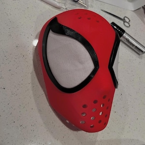 Op de afbeelding: Een rood en zwart superheldenmasker met witte mesh oogbedekkingen. Het masker heeft een reeks kleine gaatjes bij de mond. Het masker is waarschijnlijk voor cosplay of kostuumgebruik.
