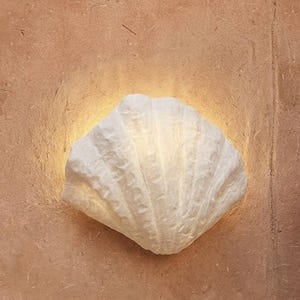 Puede incluir: Un aplique de pared blanco con textura en forma de concha marina montado sobre una superficie plana de color beige. La lámpara emite un suave resplandor dorado y cálido desde sus bordes, resaltando las crestas y curvas de la estructura.