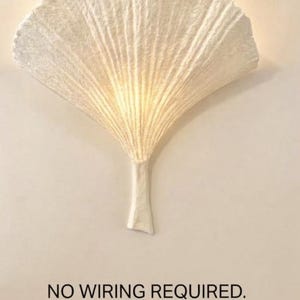 Puede incluir: Un aplique de pared blanco con forma de hoja de ginkgo y superficie texturizada. El texto dice NO WIRING REQUIRED. Includes for Puck Light Kit, con un gráfico de una luz tipo disco y un cable de carga USB mostrado debajo.