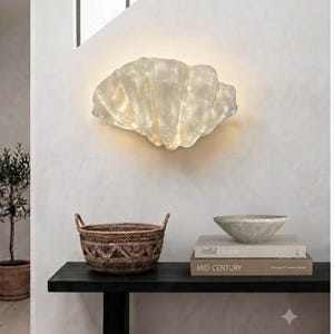 May include: A wall-mounted light fixture shaped like a large, translucent seashell with a textured, iridescent surface. The lamp emits a soft, warm glow from behind its curved, ribbed structure, creating a gentle illumination against the wall.