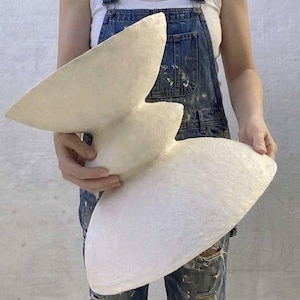 May include: A large, off-white sculptural vessel with a textured, matte surface. The object features a wide, flared top section, a narrow central waist, and a broad, rounded base, creating a distinct hourglass-like silhouette with organic curves.