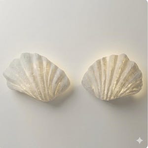 May include: Two off-white, textured wall sconces shaped like scallop shells. Each shell features a ribbed surface with a matte finish and emits a soft, warm glow from within, creating a subtle illumination against the plain, neutral background.